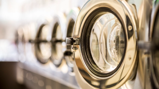 Close-up of a row of commercial washing machine doors in a laundromat, with the first chrome door open and the others receding in a blurred line.
