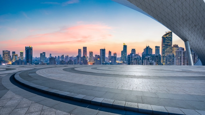 Modern city skyline with high-rises at dusk, viewed from an empty, patterned plaza. A large, curved roof element frames the upper right.