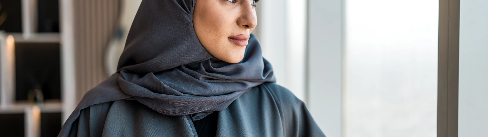 An Arabic woman wearing a gray abaya and matching hijab looks thoughtfully out of a large window with a city view.