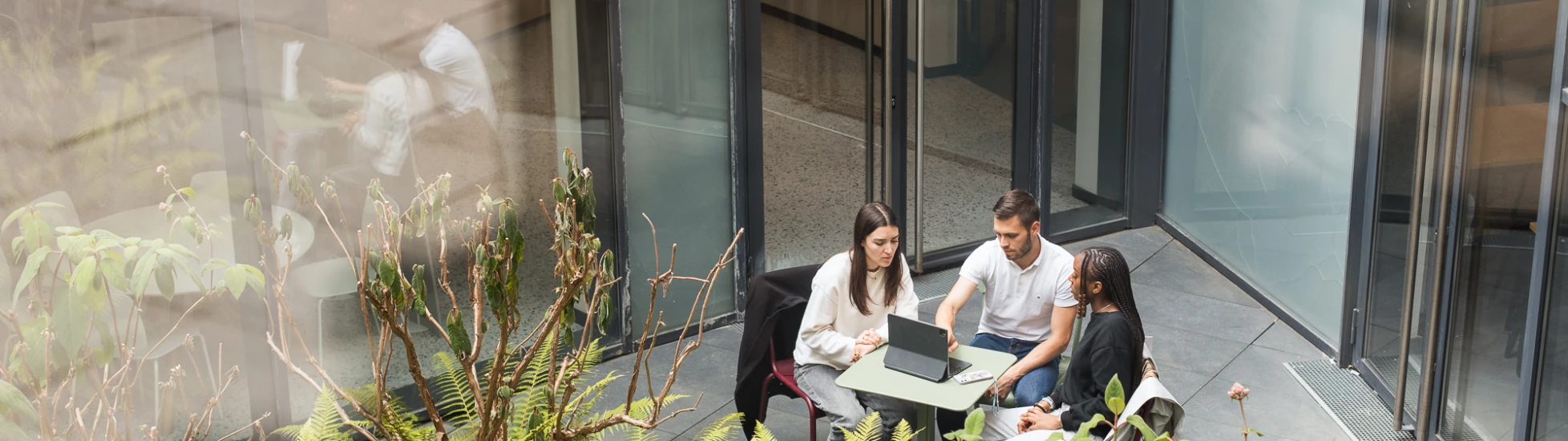 High-angle: Three people (1 man, 2 women) at small outdoor table with tablet/laptop. Glass doors behind, green plants in front.