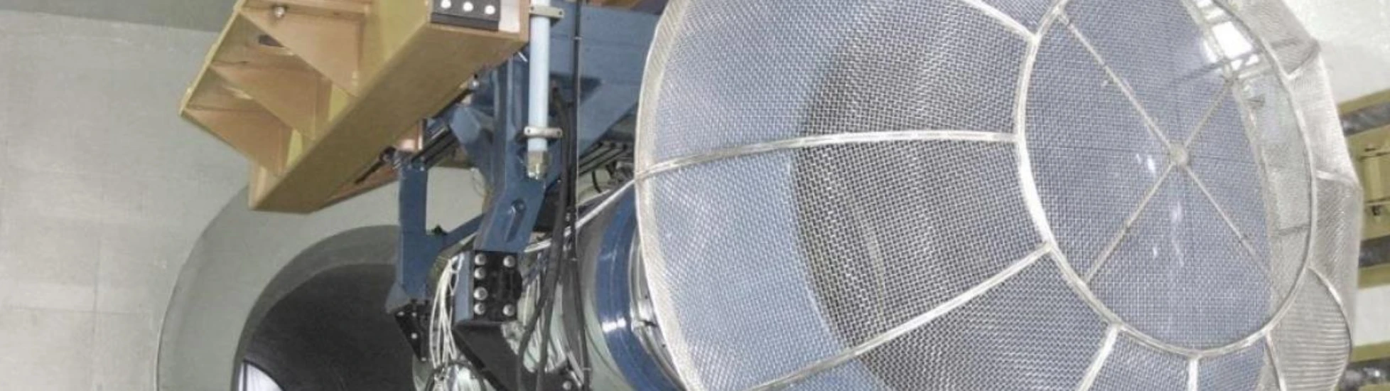 Interior view of a large, complex industrial/research apparatus: circular structure with metal mesh screen, connected to heavy machinery and a concrete tunnel exit.