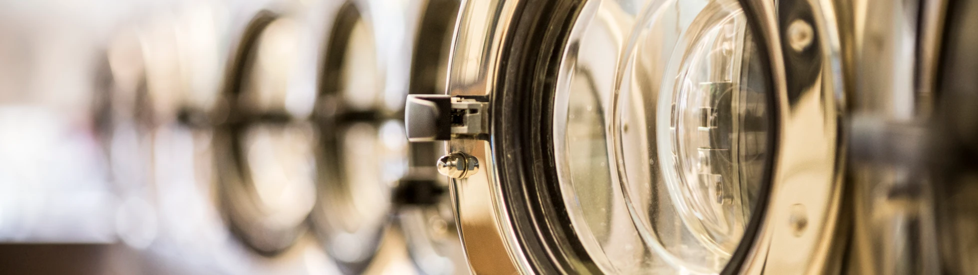  Close-up of a row of commercial washing machine doors in a laundromat, with the first chrome door open and the others receding in a blurred line.